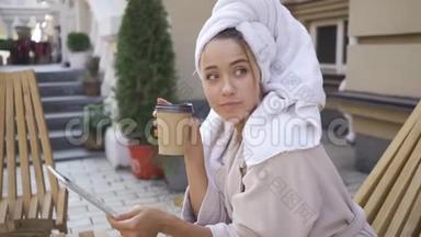 穿着浴袍、头戴毛巾、端坐在桌子旁喝咖啡、端着平板电脑的年轻可爱女子的肖像